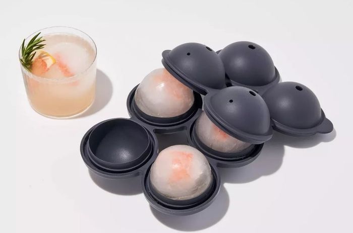 A silicone tray designed with four molds to create spherical ice cubes, with one of the cubes placed in a refreshing drink.