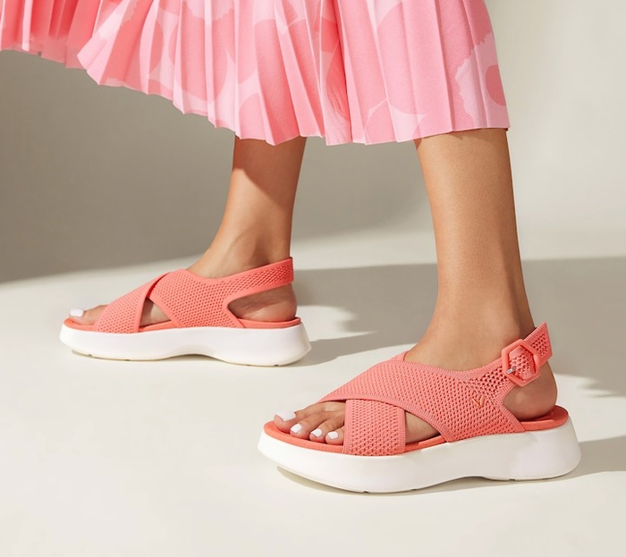 Close-up of a person’s legs in a pleated skirt, sporting stylish open-toe sandals with a thick white sole and an adjustable strap, ideal for a shopping feature