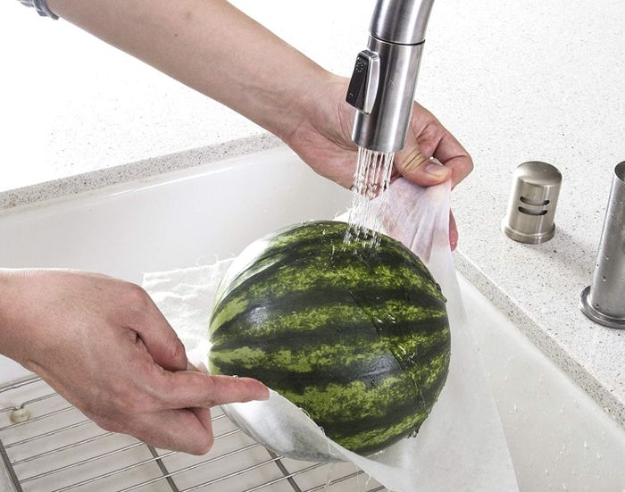 A hand holding a small watermelon, wrapped in a bamboo towel under running water