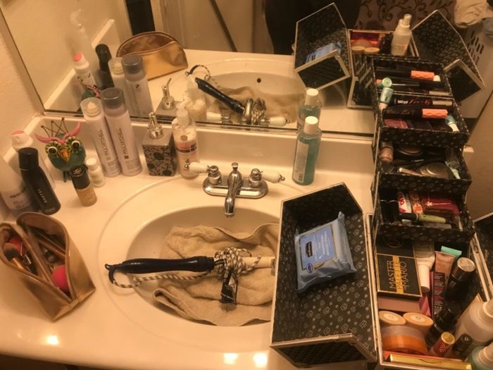 A before photo of the reviewer’s messy bathroom counter, cluttered with makeup and a curling iron in the sink