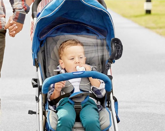 A toddler seated in a stroller with a fan attached, accompanied by adults, showcasing a portable stroller fan