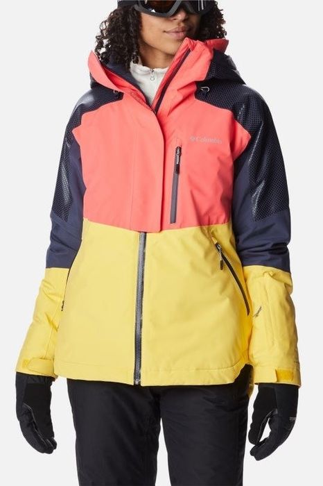 Model dressed in a color block jacket with blue, pink, and yellow panels, wearing gloves and ski goggles.