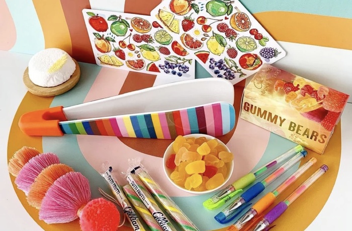 A colorful assortment of items, including gummy bears, pens, fruit stickers, brushes, a rainbow spatula, and a cupcake squishy, arranged for a shopping-themed article