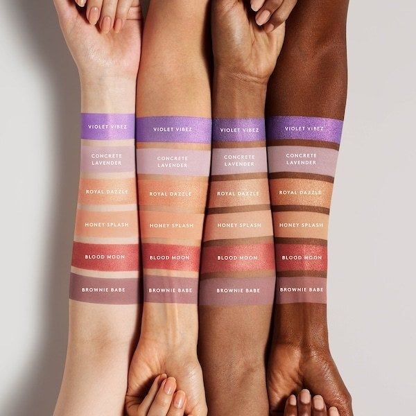 Swatches of the eyeshadow shades on arms in various skin tones. The shades include a sparkling purple, a lavender, matte and glittery peach shades, a glittery red, and a mauve.