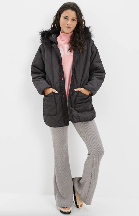 Model wearing a black quilted jacket that falls below the waist, hands tucked into the pockets
