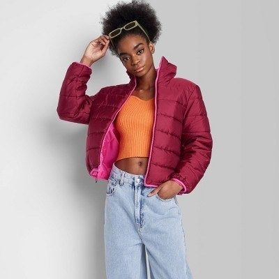 Model rocking a dark pink, waist-length puffer jacket, one hand holding sunglasses and the other resting in their pocket