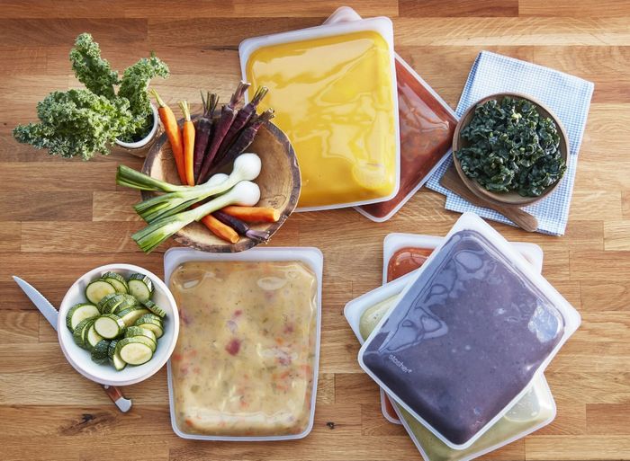 Reusable silicone food storage bags placed on a wooden surface with an assortment of vegetables and prepared meals.