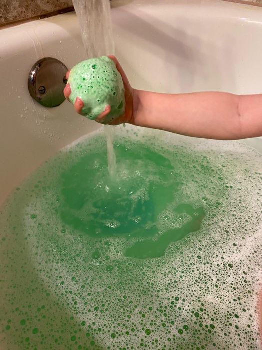 A hand holds a green bath bomb under running water in a bathtub, creating green foam and bubbles in the water.