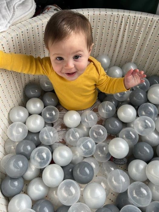 A baby smiles up at the camera, sitting in a playpen surrounded by white and gray plastic balls, wearing a long-sleeve top.