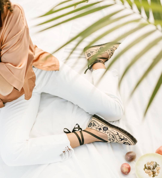 A model showcasing the tan closed-toe espadrilles with black illustrations covering them, featuring a black sole and black laces wrapped around the ankles.