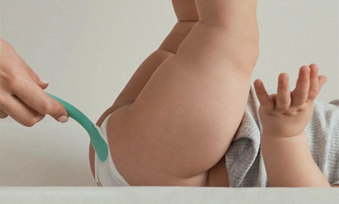 A baby rests peacefully as a soft, flexible brush gently applies cream to their skin, with the text reading,