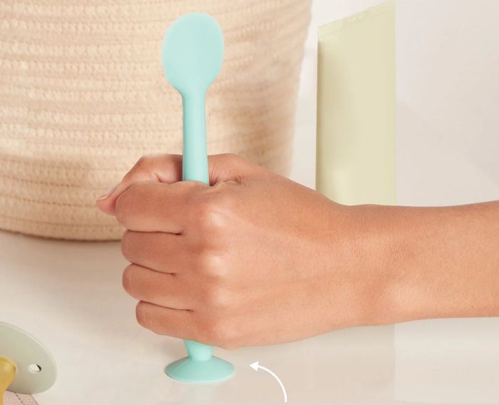 A person holds a spoon with a suction base, showcasing its ability to stay put beside a bag and tube for added convenience.