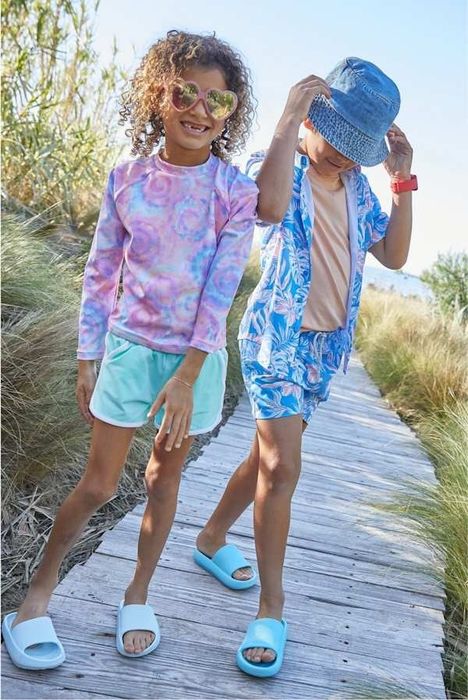 Kids enjoying summer on a boardwalk. One child has curly hair, heart-shaped sunglasses, a tie-dye long-sleeve, and shorts. The other wears a patterned shirt, shorts, and a hat.