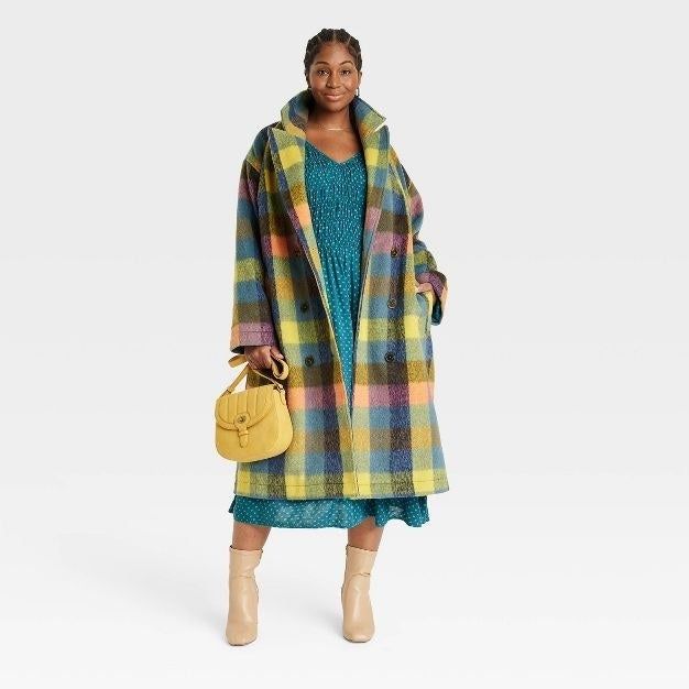 Model showcasing a below-knee length coat in a vibrant yellow, green, blue, and pink plaid, hands tucked into pockets