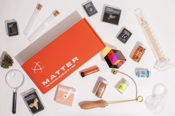 "A collection of science and nature items such as minerals, a magnifying glass, test tubes, and a beaker arranged around an orange 'Matter - The Science Museum in a Box' kit."