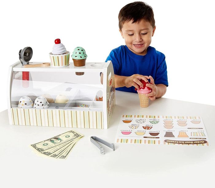 A child model placing a topping on an ice cream cone, while the stand, menu, and pretend money rest on the table