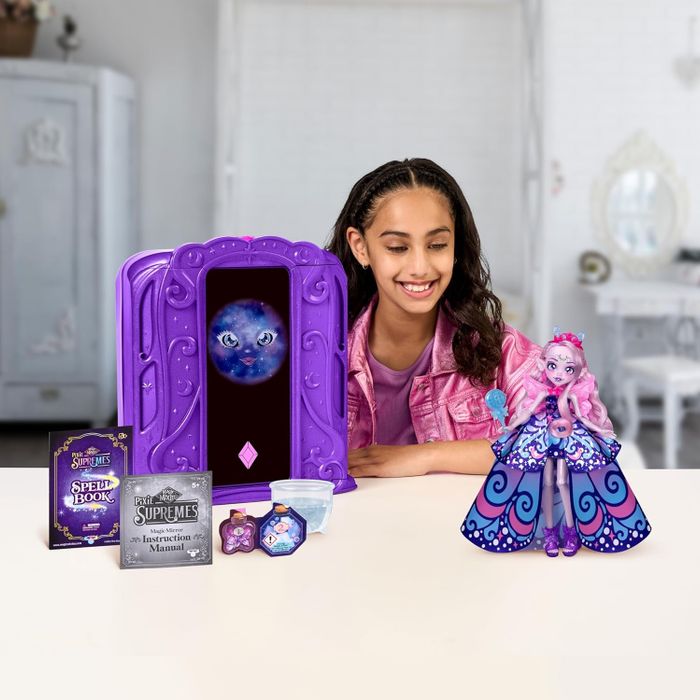 A girl with a magic-themed toy set, complete with a spell book, instruction manual, digital screen, and a doll in a patterned dress with wings