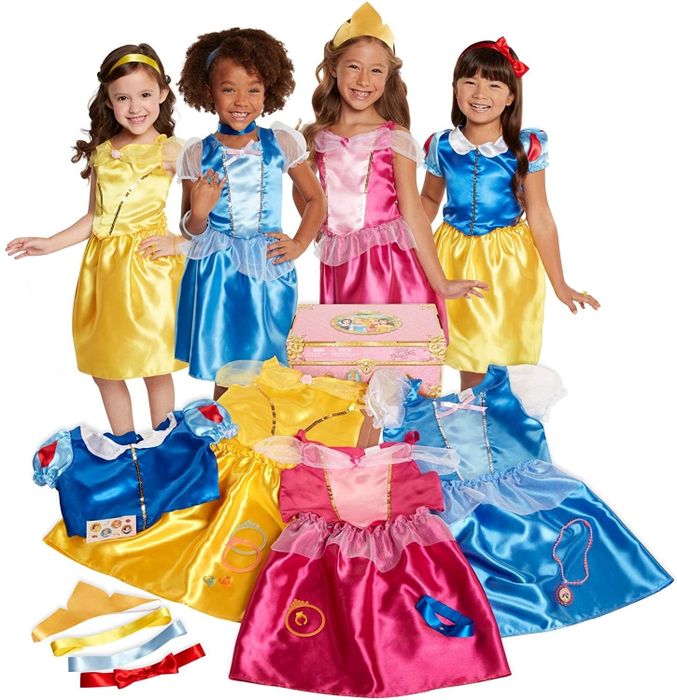 Four children dressed as princesses, posing beside a pink trunk filled with yellow, pink, and blue satin gowns.