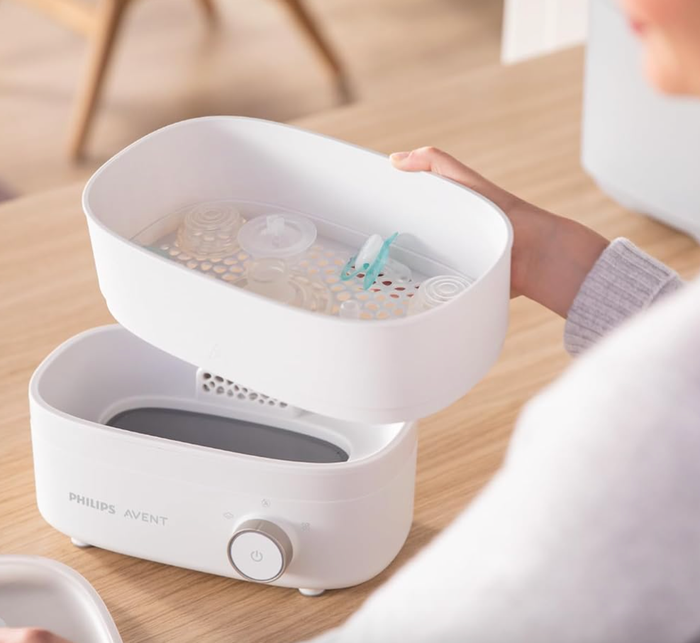 A person using a Philips Avent bottle sterilizer on a wooden table, holding the lid open to reveal the compartments inside.