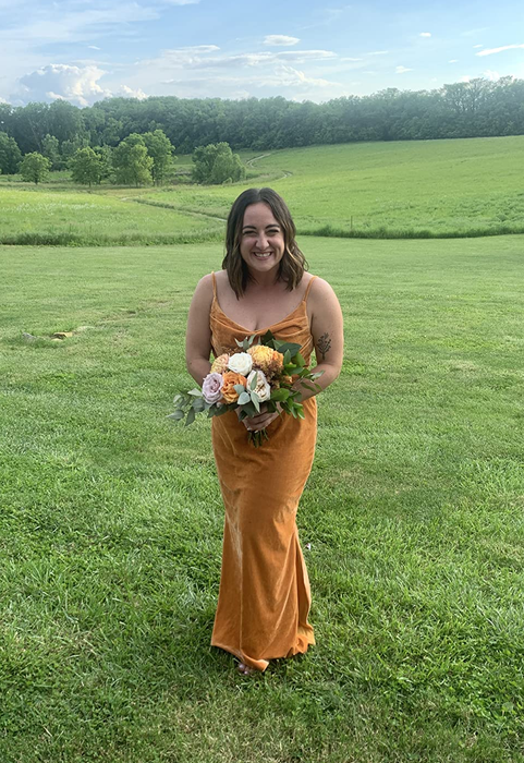 A reviewer wearing the mustard-colored dress, holding a bridesmaid bouquet