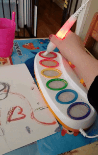 A hand using a glowing pen to color on a child’s artwork placed on an interactive coloring pad with vibrant circles