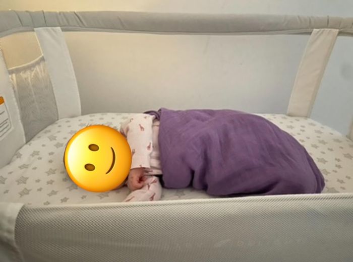 A baby peacefully sleeps in a crib, wrapped in a blanket with a smiley face emoji covering their face.