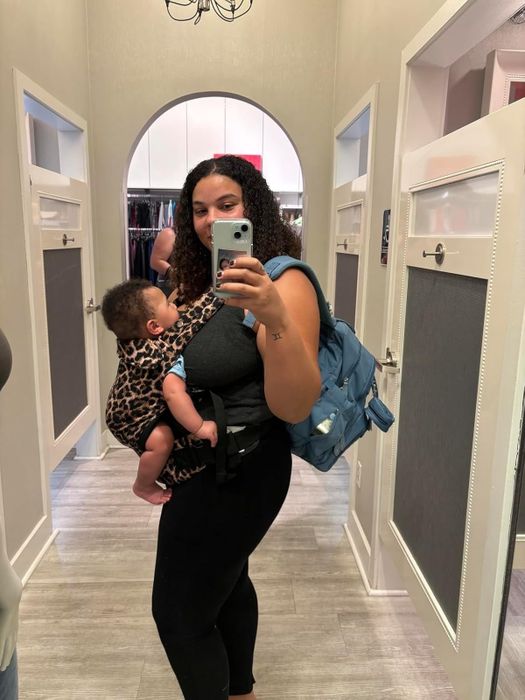 A woman takes a mirror selfie while cradling her baby in a leopard print wrap. She’s dressed in casual clothes and carrying a backpack, while shopping in a store.