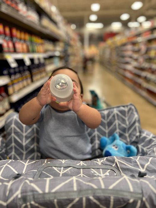 A baby sits in a grocery store shopping cart holding a bottle, surrounded by shelves filled with various products in the background.