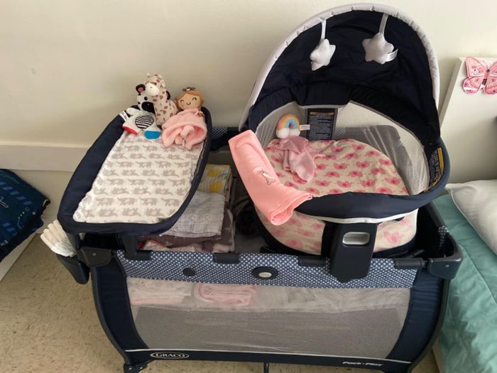 A Graco Pack 'n Play playard is set up with a bassinet insert. Inside, there's a plush blanket, a swaddle, a pacifier, and several stuffed toys, making it a cozy spot for a baby.