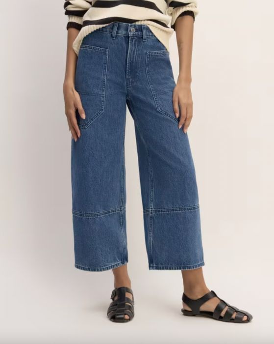 A person standing in cropped denim pants paired with black sandals, with a cream top partially visible. The face is not shown.
