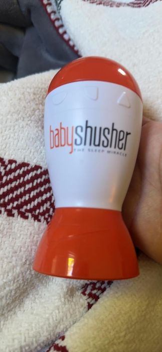 A hand holds a Baby Shusher, a white and red device designed to aid infant sleep with soothing sounds.