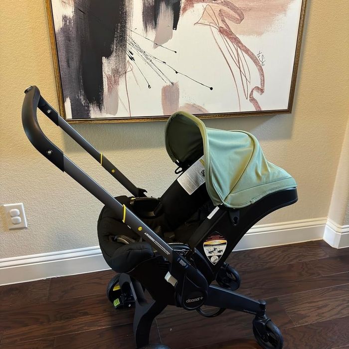A compact black stroller with a green canopy is placed indoors, leaning against a wall beneath an abstract painting. There are no people visible in the scene.