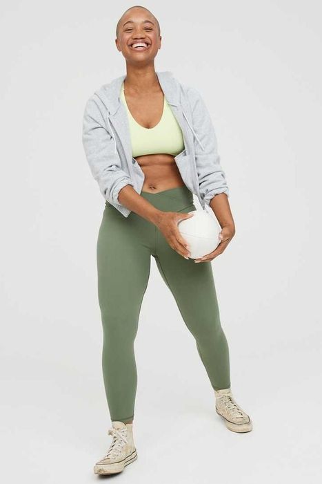 A model wearing green leggings with a crossover waist, paired with tennis shoes and a zip-up hoodie