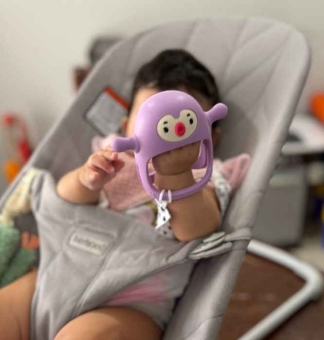 A baby enjoys sitting in a bouncer while holding a purple toy with a face, surrounded by various other toys in the background.