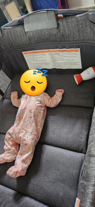 A baby sleeps peacefully in a printed onesie in a crib, with a bottle lying beside them. The crib includes text with details about usage and safety instructions in both English and Spanish.