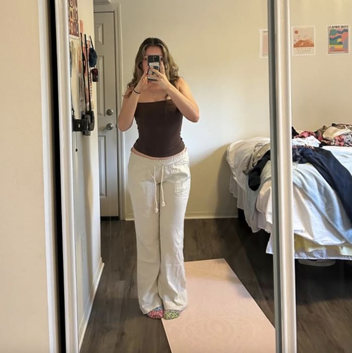 A person taking a photo of themselves in the mirror with their phone, wearing a tank top and white pants, with a messy bed in the background.