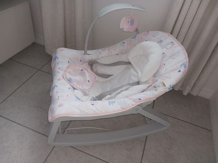 A baby bouncer seat featuring cushioned head support and secure safety straps. Suspended above is a mobile with two soft elephant toys.