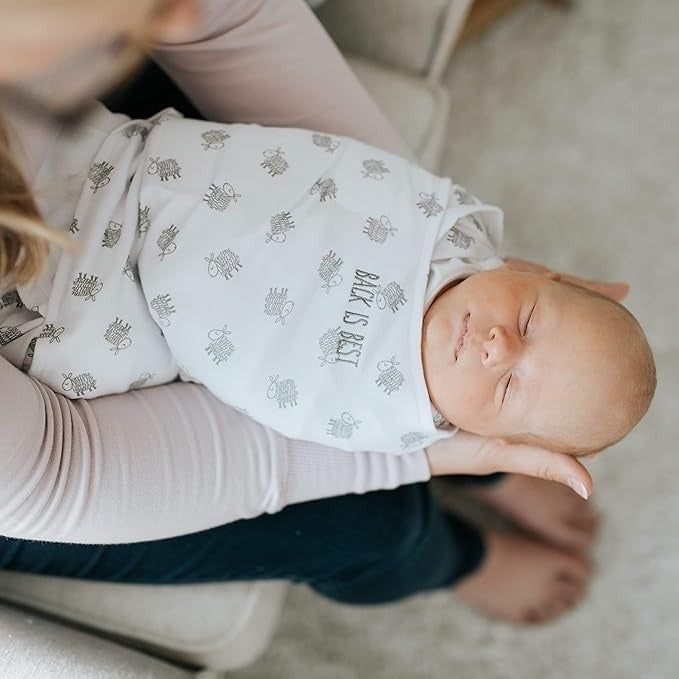 A baby is swaddled in a blanket with the words 'Babe is Best' printed on it, held by an unseen adult in a light shirt and dark pants, sitting on a beige carpet.