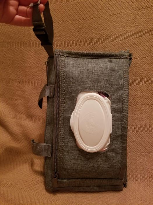 A hand holds a compact, rectangular fabric bag with a zippered pocket and an integrated white plastic wipe dispenser.