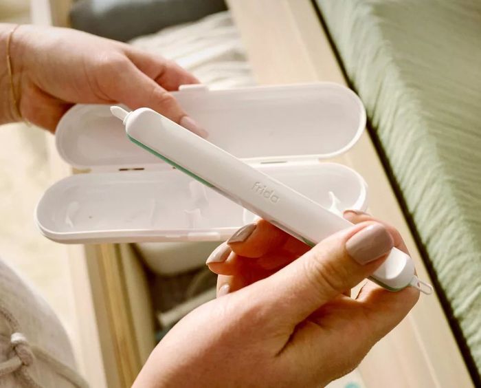 A person holds the FridaBaby cleaning tool with its case, demonstrating how easy it is to use. Text on the case reads: