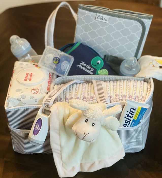 A baby gift basket packed with essentials like diapers, wipes, a baby bottle, pacifiers, Desitin cream, a soft sheep plush toy, a bib with 'Ribbit Ribbit' on it, and handy storage pouches.