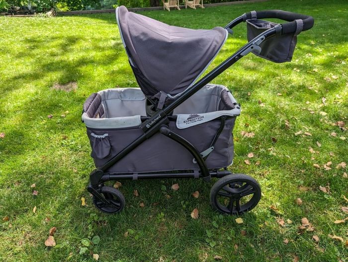A gray baby stroller with a canopy, large wheels, and plenty of storage space, parked on a grassy patch.
