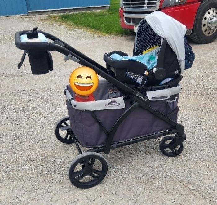 A Wonderfold stroller sits on a gravel path, surrounded by various bags and items, with a large smiling emoji covering the face of a child inside.