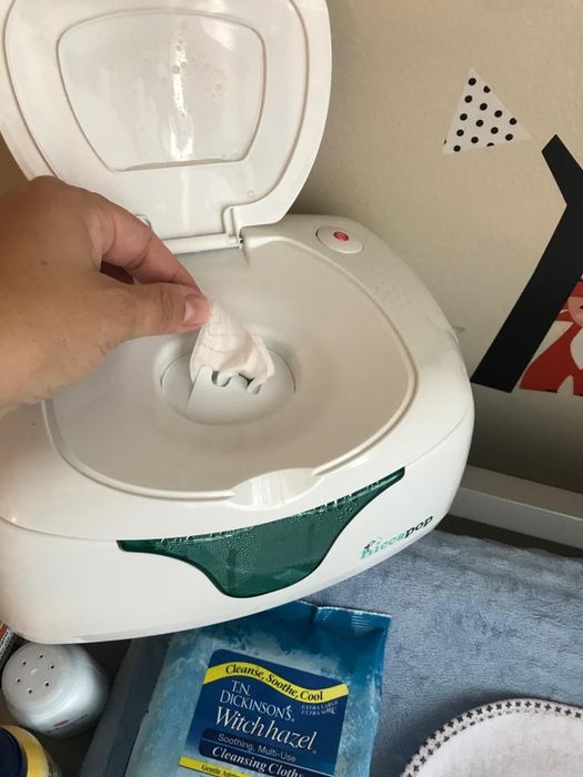 A person removes a baby wipe from a white wipe warmer, with a package of Dickinson's witch hazel cleansing cloths visible on a gray surface nearby.