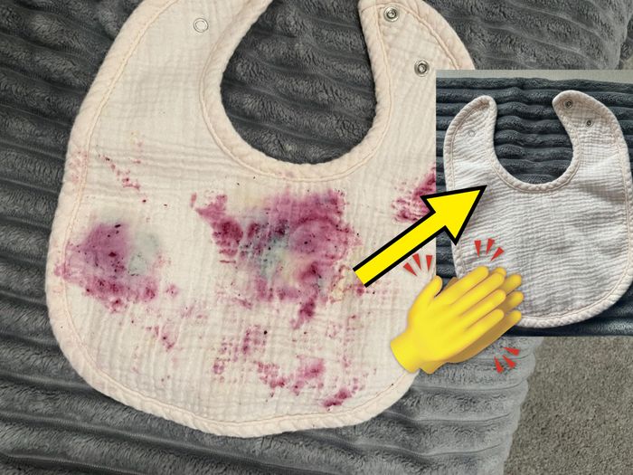 A white baby bib with purple food stains rests on a textured gray surface, waiting to be treated.