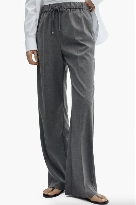 A person models gray wide-leg pants and a white shirt, standing in profile with hands casually tucked into pockets.