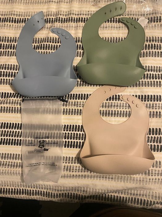 Three silicone baby bibs with pockets, laid out on a patterned surface: one blue, one green, and one beige. Each bib features adjustable snap closures.
