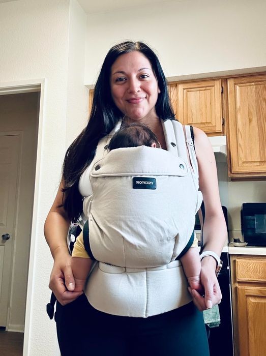 A woman beams with a smile while holding her baby in a MamKangaroo baby carrier. They are standing together in a cozy kitchen.