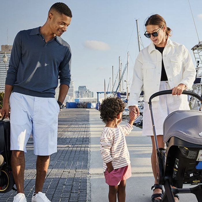 A family browses stroller options by a marina: a man in a long-sleeve shirt and shorts, a woman in a white jacket and shorts, accompanied by a child.