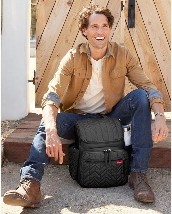 A man wearing a casual tan jacket, a white shirt, and jeans smiles while sitting outdoors next to a modern black backpack. This is part of a shopping category article.